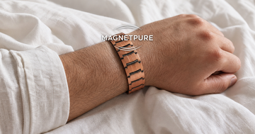 Man's forearm wearing a copper magnetic bracelet resting on white bedsheets in soft morning light